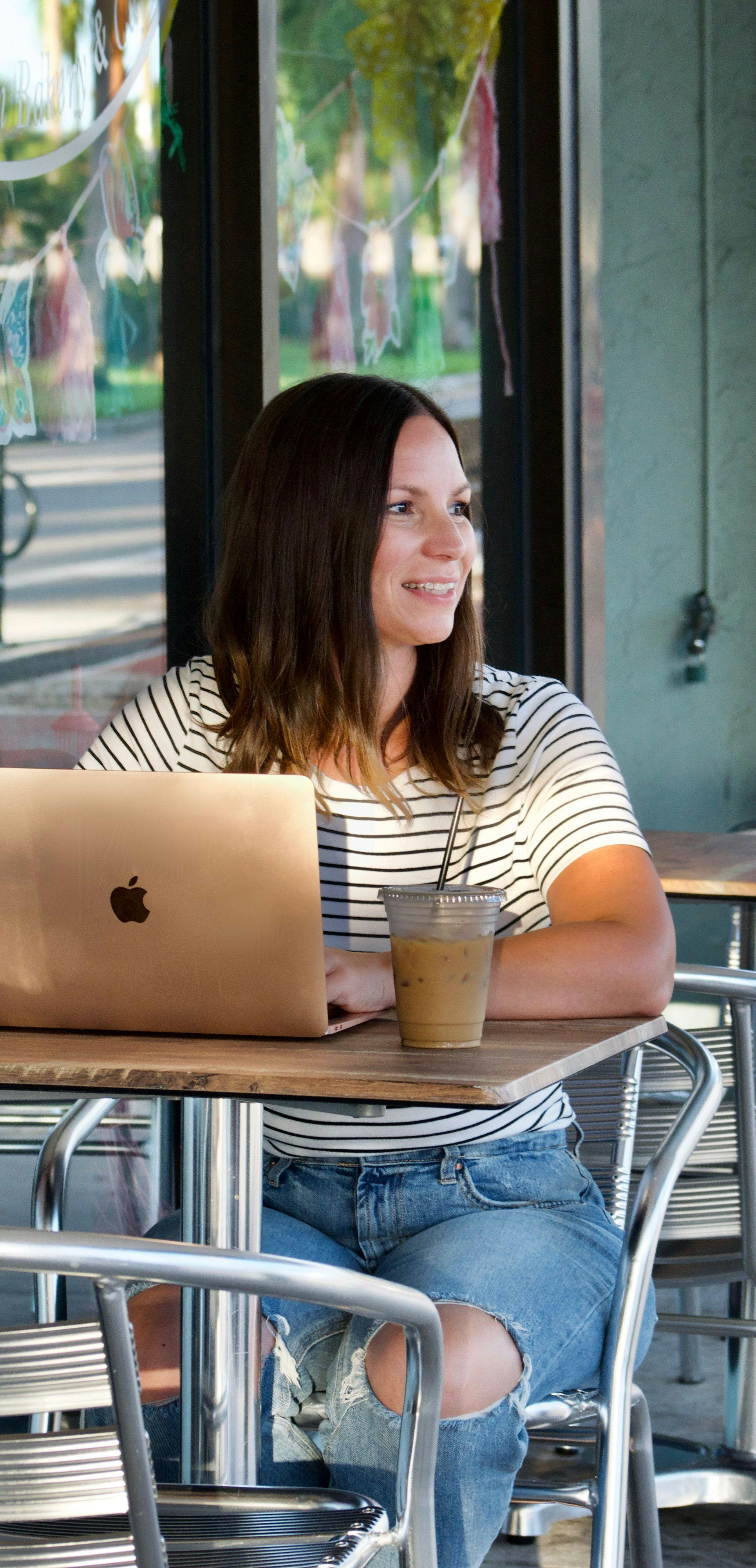 Leigh Smith, e-commerce copywriter of product descriptions of Leigh Smith Copy sits outside of a cafe on a computer, smiling with coffee.
